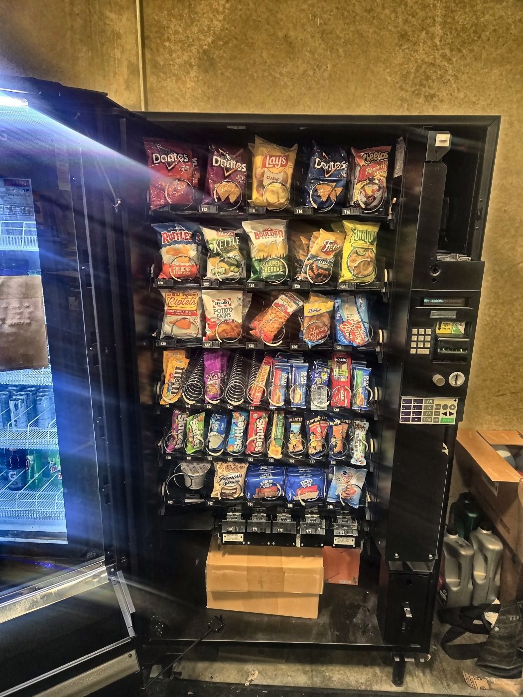 A snack machine next to a lit cold drinks case stocked with bottles and cans.