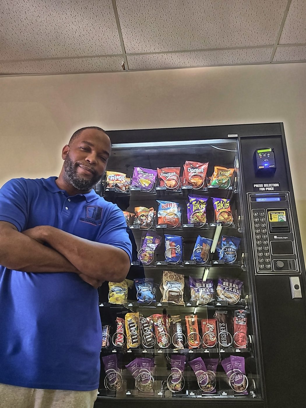 The owner of SKS Vending standing with his arms folded beside one of his fully stocked machines, wearing a blue SKS Vending polo shirt.
