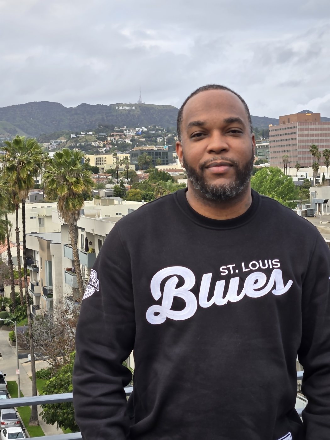 The owner of SKS Vending in a St. Louis Blues sweatshirt, photographed on a balcony with the Hollywood sign on the hills behind him.